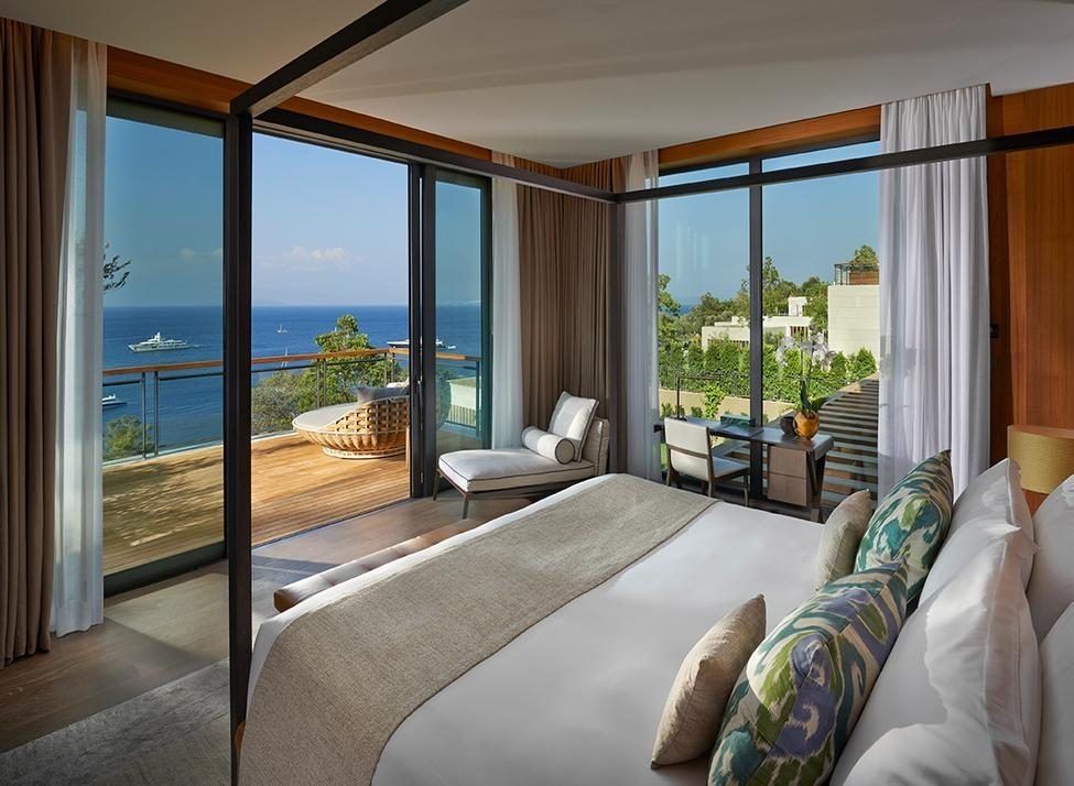 A double bed in the foreground looks out onto a wraparound terrace with round lounge chair and a balcony, with the sea, olive trees and yachts visible.