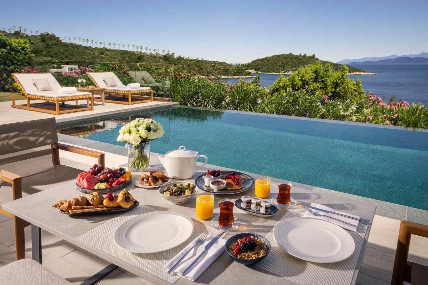 A table laid with breakfast of fruits and pastries, overlooking an infinity pool and with views over the bay and sea.