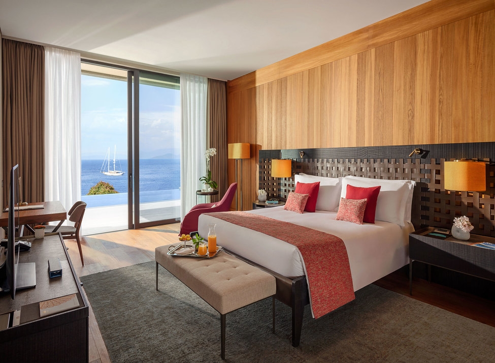 A double bed with white sheets and red cushions and throw, with a wooden headboard and footstool laid with a breakfast tray. Sliding glass doors frame views of the sea and a yacht in the distance.