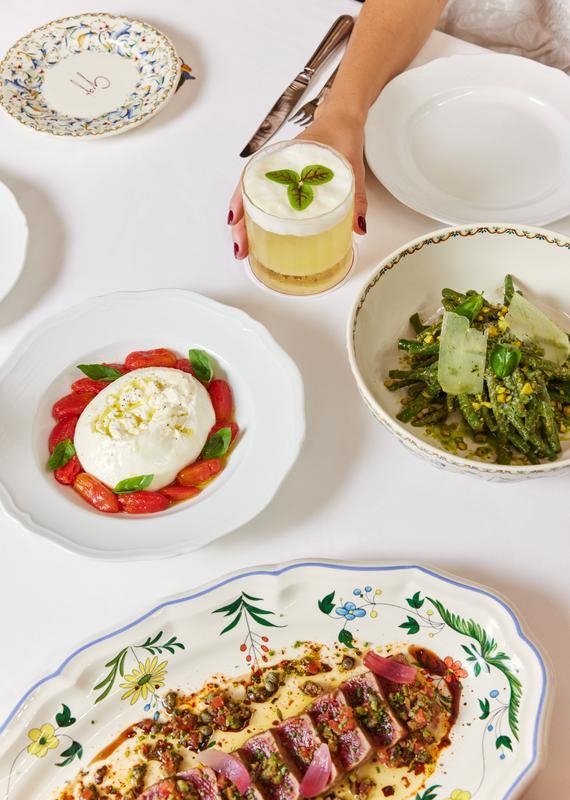 An overhead shot of a white tablecloth laid with Mediterranean dishes, and a woman's hand holding a cocktail