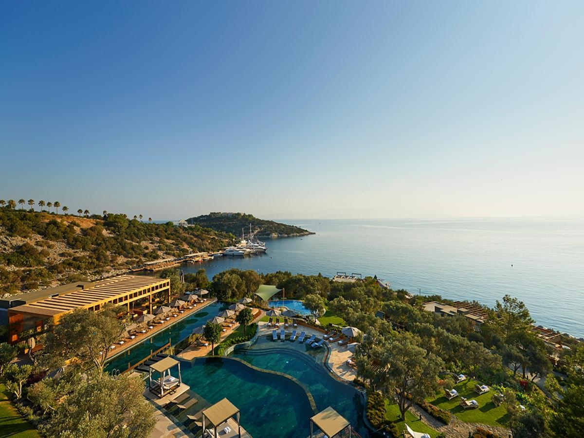 An aerial image of a coastal resort in Bodrum, Turkey, showing several pools, beach cabanas and a covered outdoor restaurant, with gardens and olive trees leading to the sea