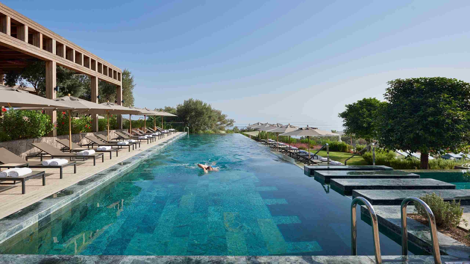 A long infinity pool with a person swimming front crawl. A row of sunbeds lines the edge.