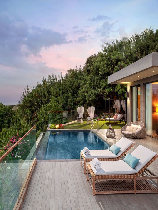 Two sunbeds with cushions and towels, in front of a small infinity pool with a small garden and trees on the other side and a partial views of a suite behind.