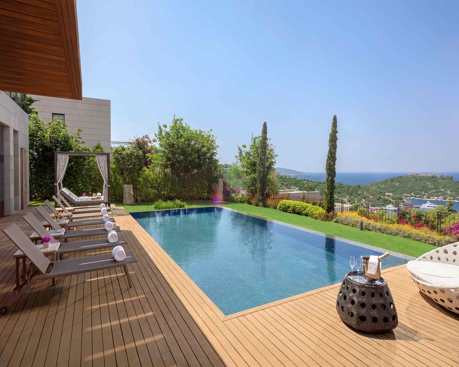 A wooden deck with infinity pool and a row of sun loungers with one cabana. There's a building behind and the pool looks out to views of the bay and coastline beyond.