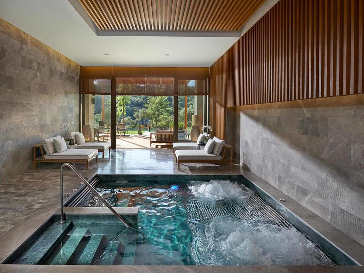 An indoor spa hydrotherapy pool with steps, bubbling water and four lounge beds behind, with floor to ceiling glass doors leading out to gardens with trees and more loungers.