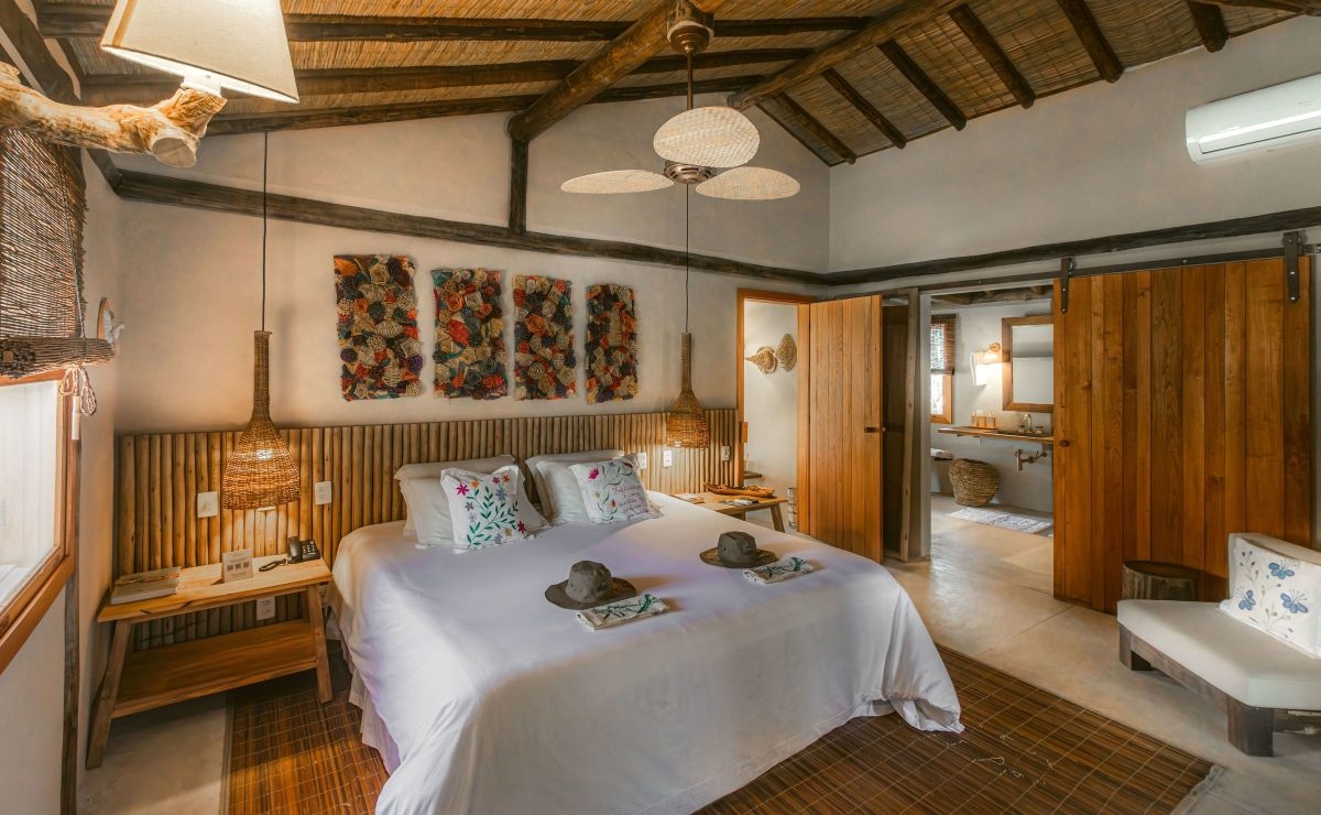 A softly decorated master suite with colourful abstract art on the wall and a wooden headboard in front of a king-size bed with white linen and two hats laid out for guests to use. Rattan lampshades hang gtom the ceiling and a wooden sliding door leads to the ensuite bathroom.