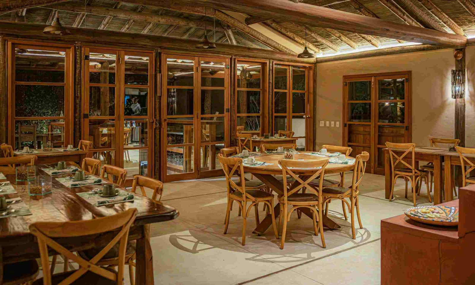 A restaurant at night, with tables set inside under a thatched roof and tall doors with wooden frames leading to the outside deck with more tables.