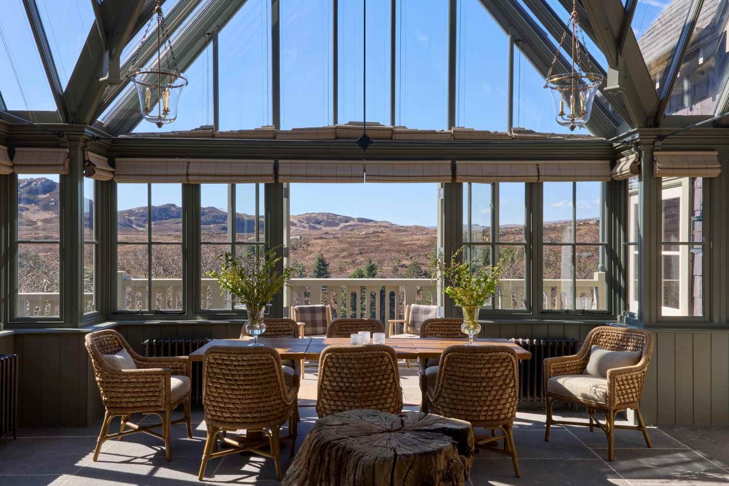A large dining table set with eight rattan chairs, with two vases. A glass roof and huge windows frame views of the surrounding wilderness.