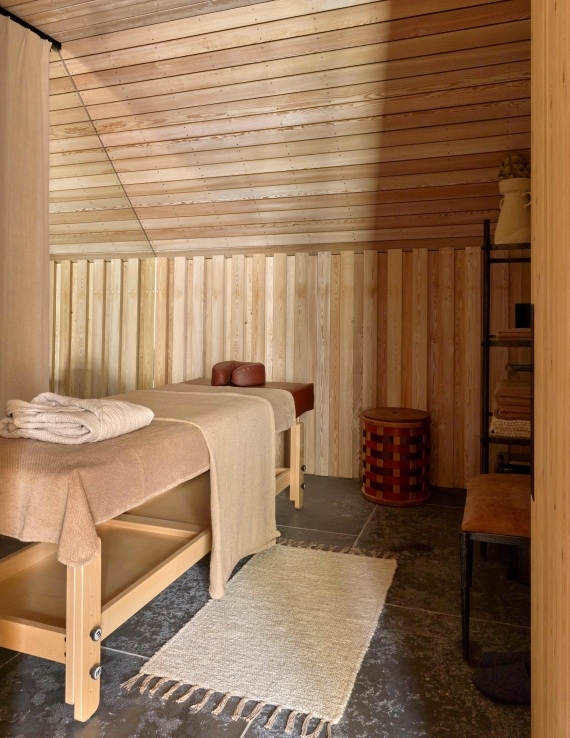 A wooden massage table set for one person, with a brown headrest, in a treatment room with wood-panelled walls, dark grey floor tiles and a pale rug.