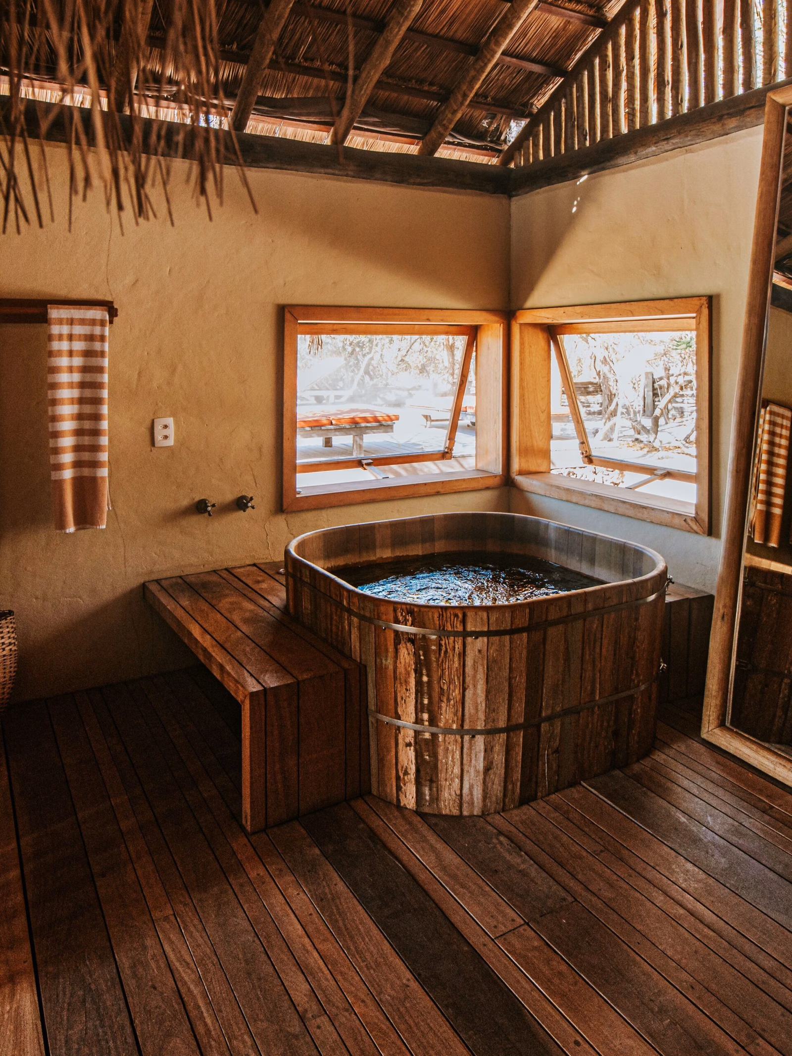 A wooden hot tub with a bench and wooden floors in the same dark colour, in front of two windows and below a thatched roof.