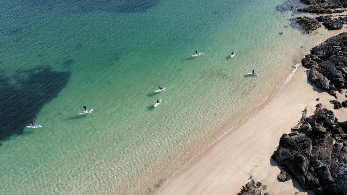An aerial view of clear, green-tinted water with several people on stand-up paddleboards, just off the coastline with a pale sand beach and rocks.