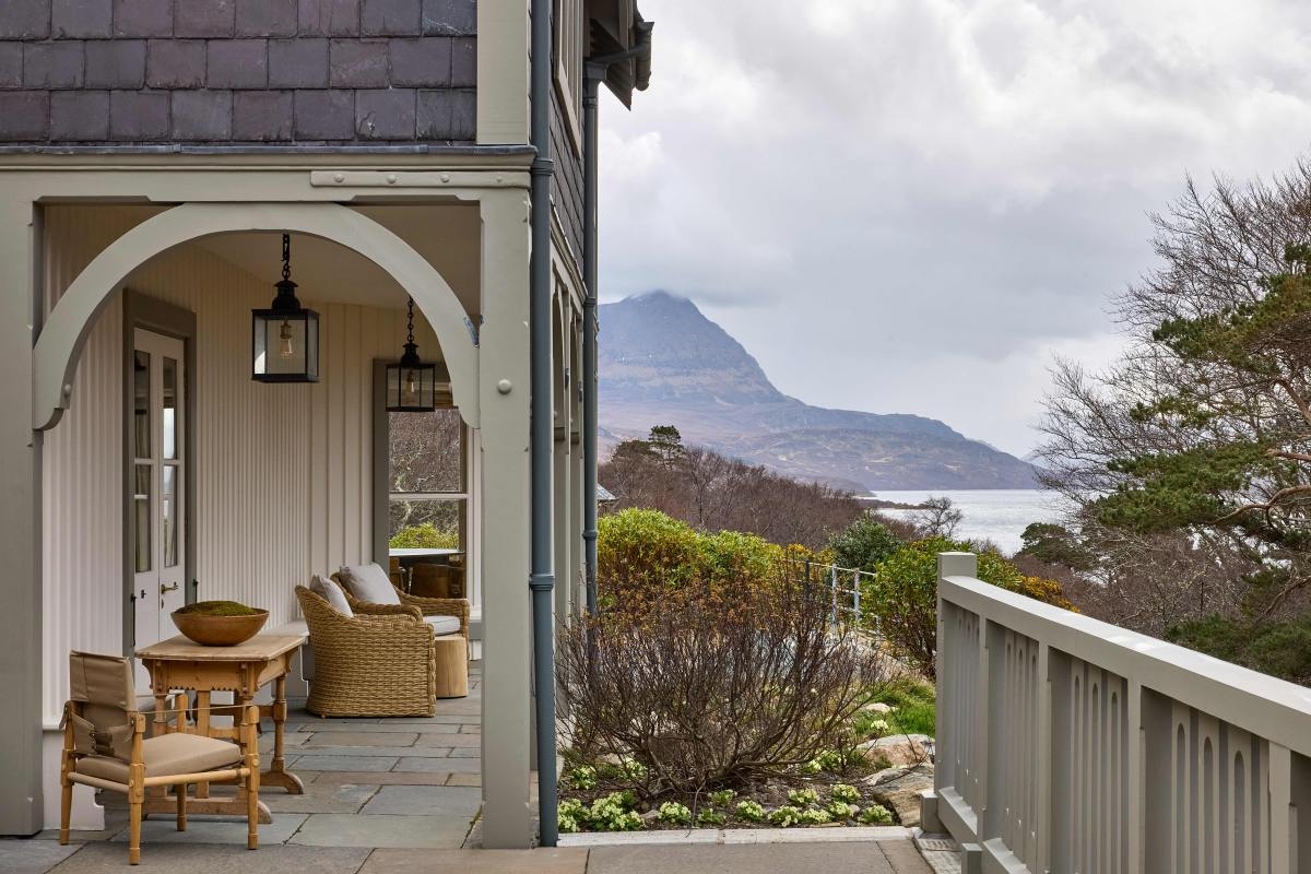 A covered verandah has rattan furniture with cushions and a white door, and looks out to views of a loch and Ben Hope mountain.