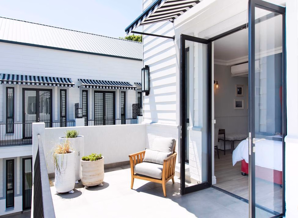 Glass doors opening out from a hotel suite to a large terrace with low stone walls, three potted plants and a chair with cushions. A bed is visible inside, and the terrace looks out to other hotel buildings, painted white with black and white awnings.