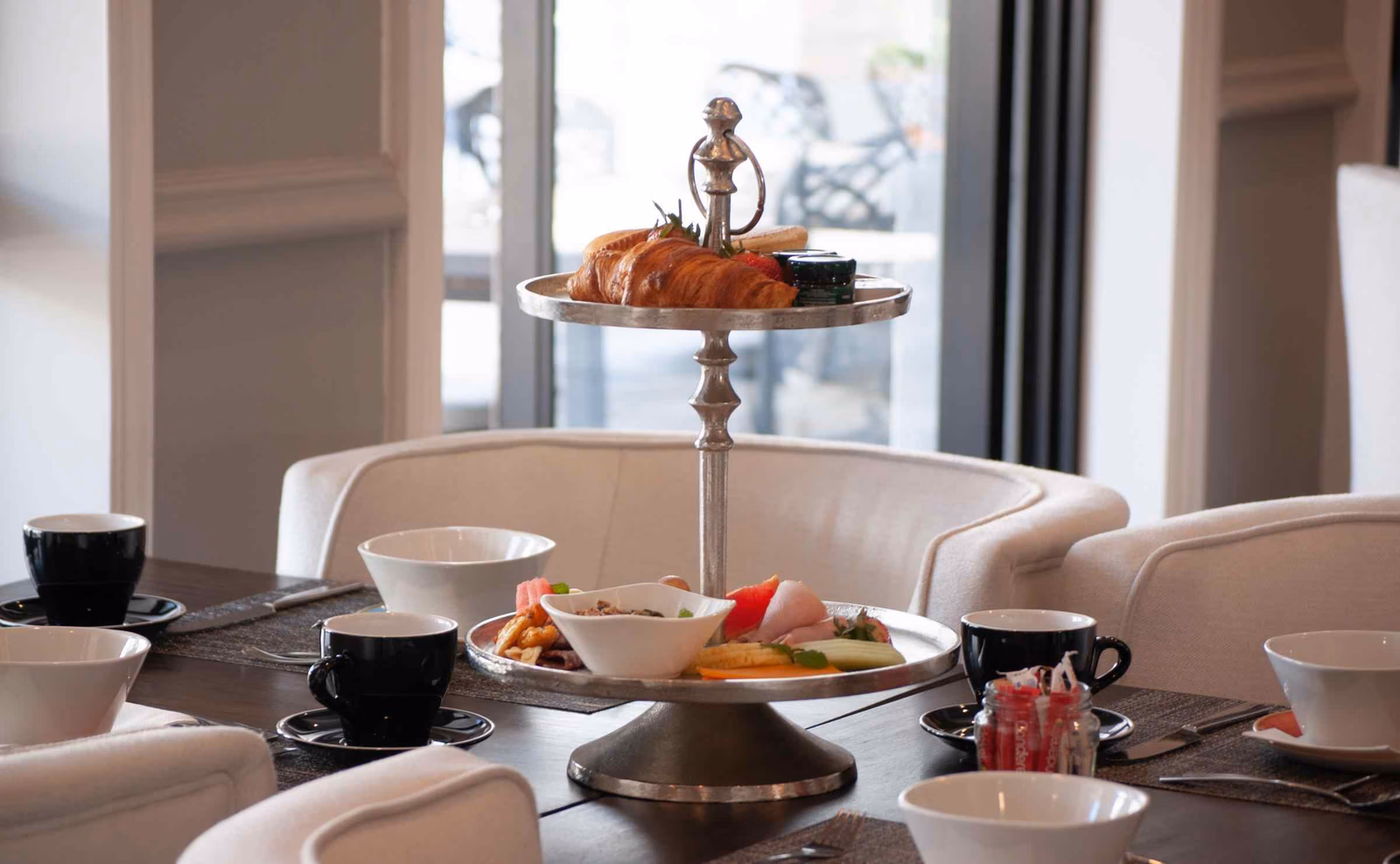 A close-up view of a table set for breakfast, with cream armchairs, cups and saucers and a cake stand with croissants, fruit and jams. Glass doors in the background lead to a terrace.