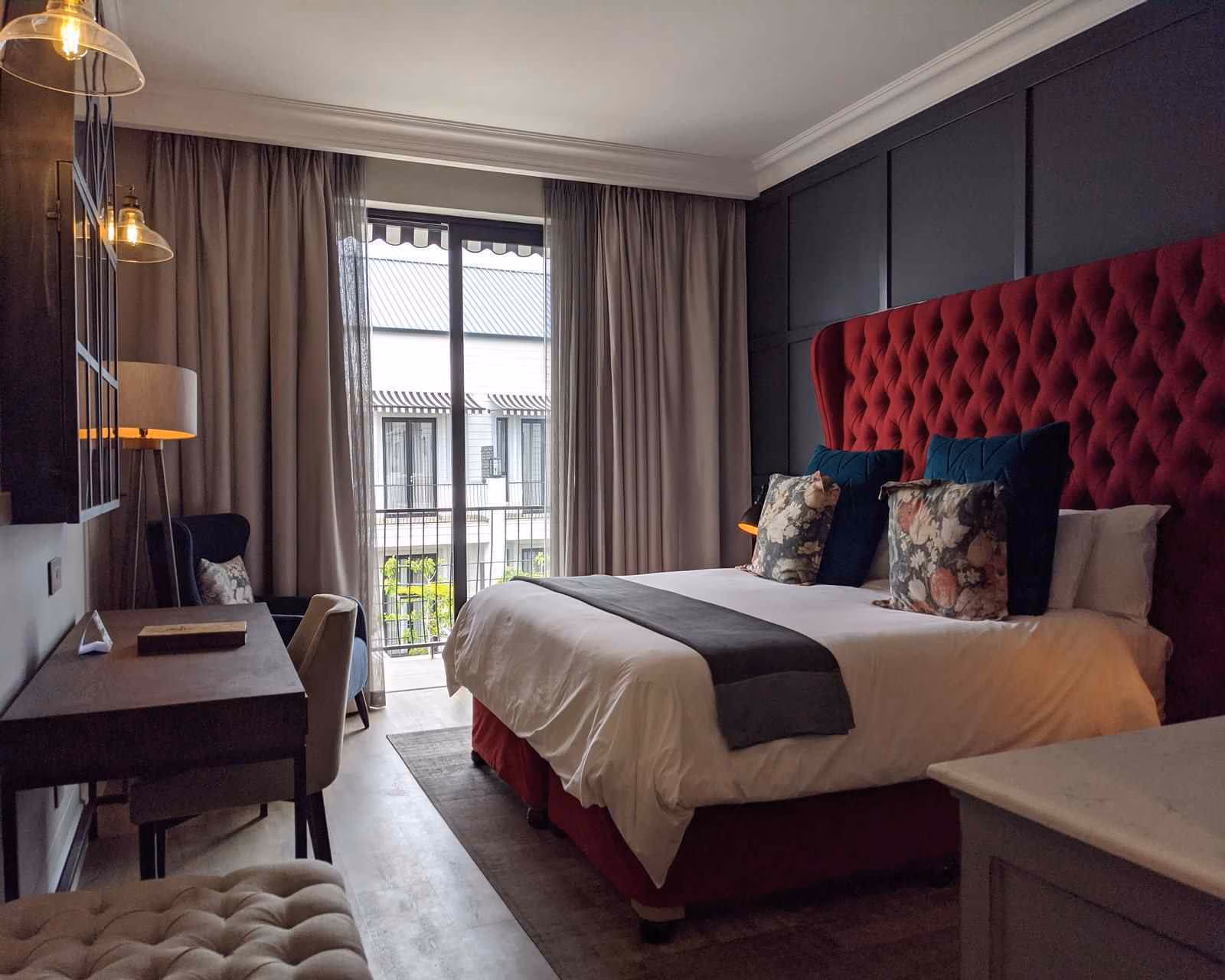 A dimly lit hotel bedroom with red headboard and partly drawn curtains showing views across a courtyard to a white building. There's a double bed, several lamps and a wooden desk with chair.