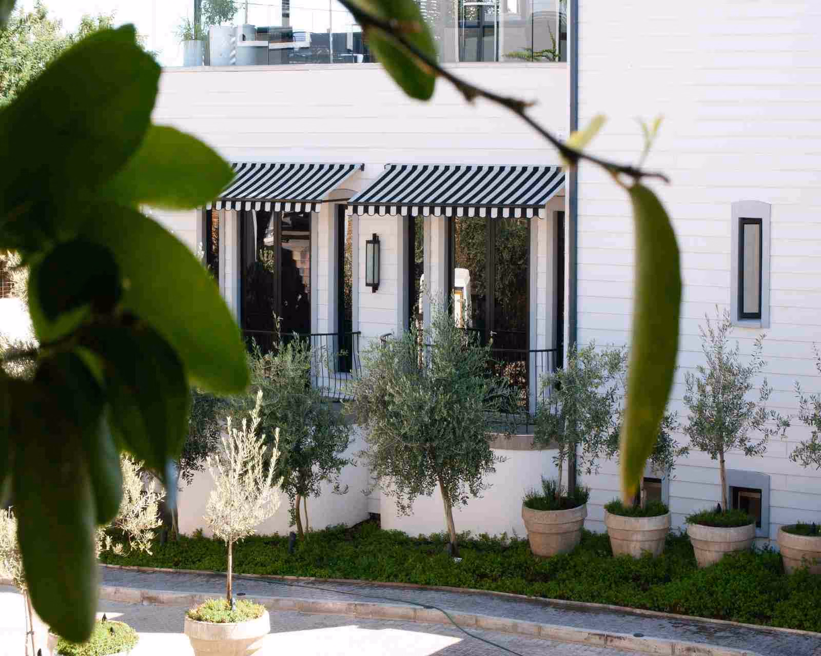Exterior view of a white, timber hotel building with black-and-white awnings and iron balconies with trees and potted plants outside.