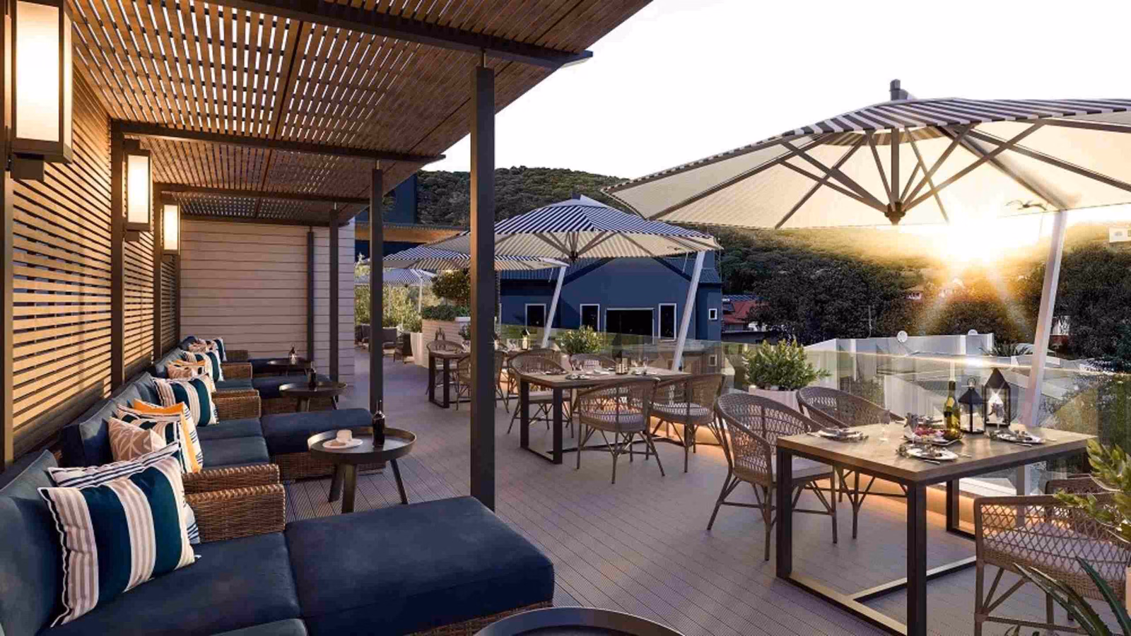 An open-air terrace with a pergola over a row of navy sofas with patterned cushions, with a parallel row of tables and chairs with large black and white parasols, looking out to sunset views.