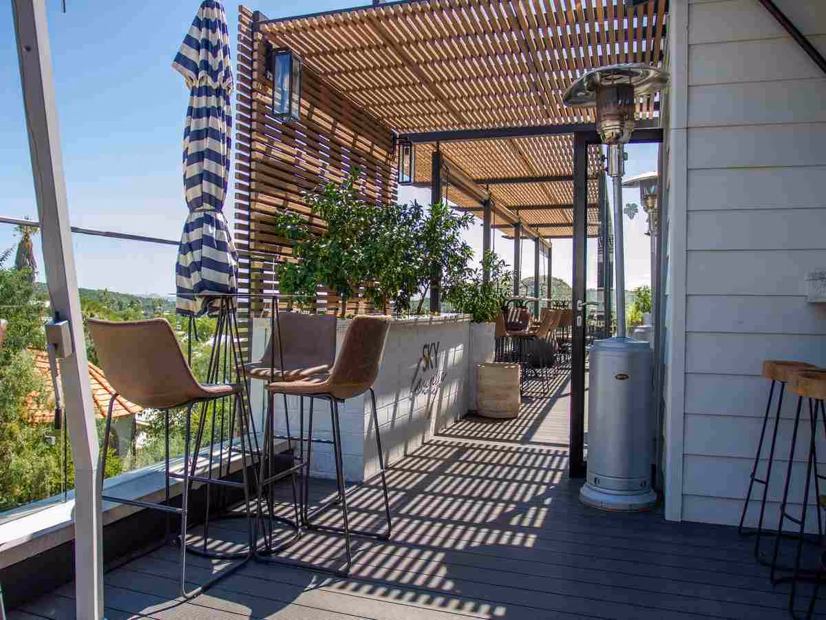 A rooftop terrace with pergola, bar stools, patio heaters and striped parasols, with views of greenery visible all around.