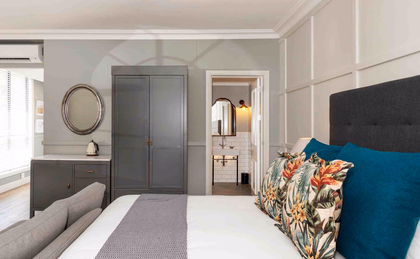 A double bed with white linens and bold patterned cushions, with a dark grey wardrobe and cupboards and a door looking into an ensuite bathroom with white tiles and a mirror.