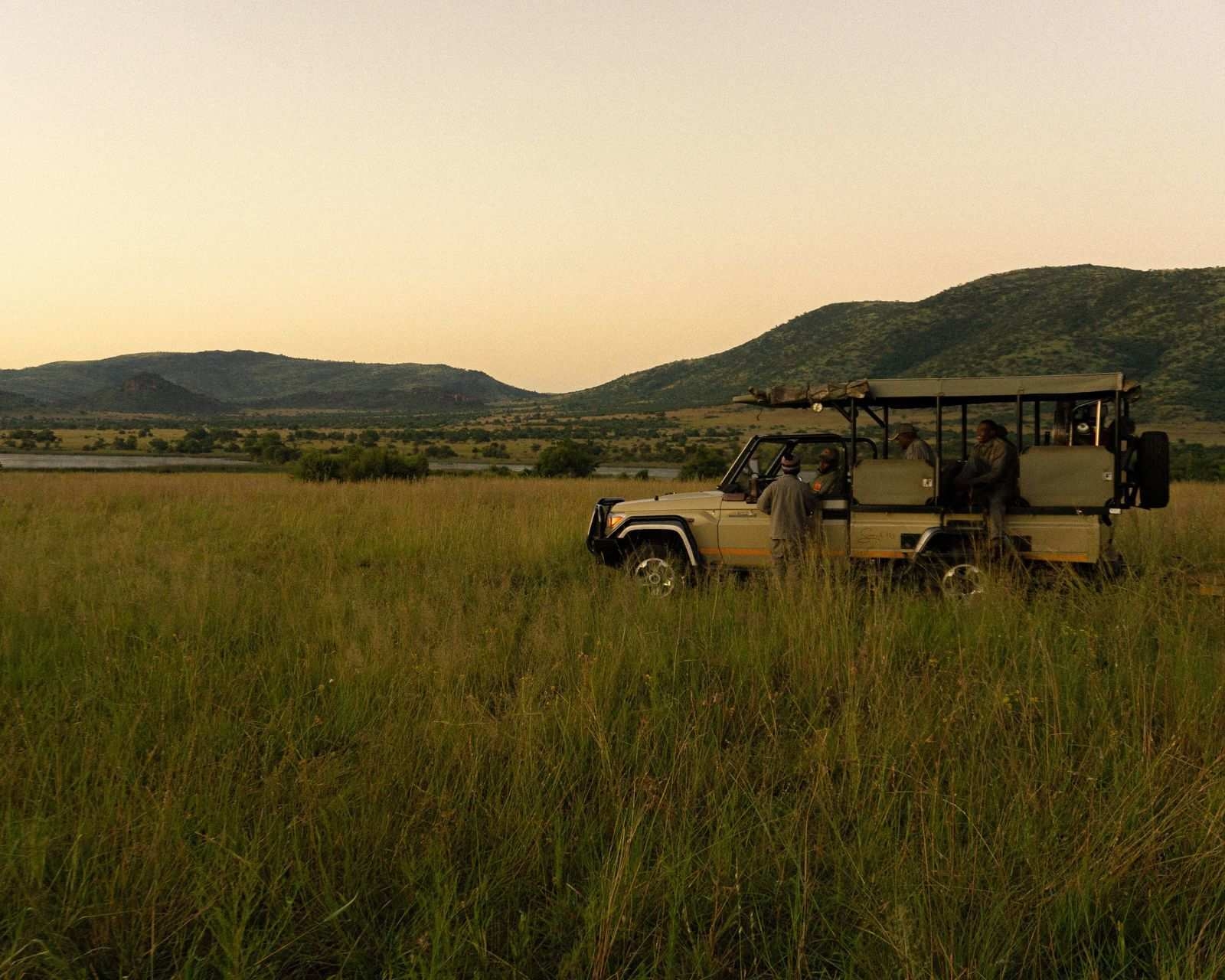 An open-sided safari vehicle parked in tall green grass near a river, with a guide standing next to it and passengers seated inside