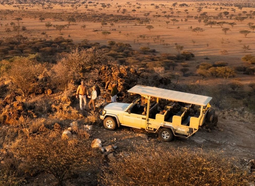 Aerial view of a yellow open-sided safari vehicle parked near a rocky outcrop in a vast savannah, with a small group of people standing outside