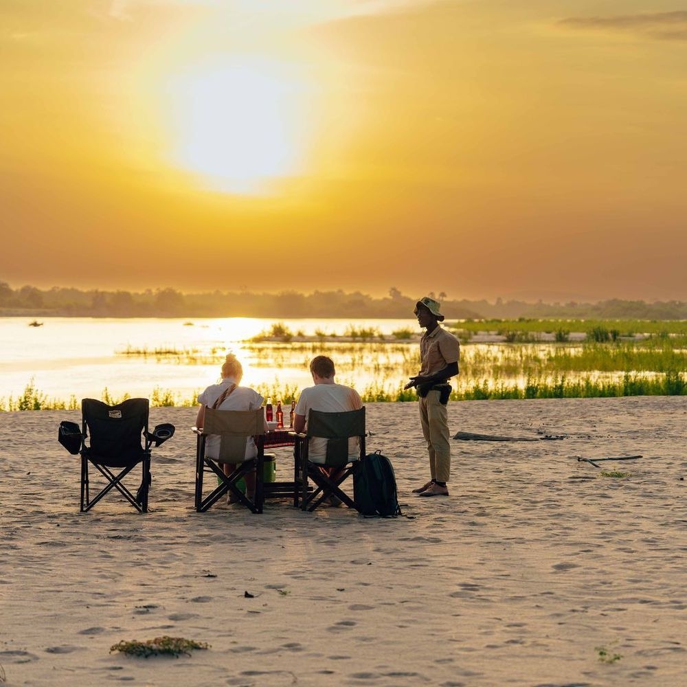 A couple sitting in camp chairs on a sandy riverbank, enjoying drinks at sunset while a safari guide stands nearby in the Okavango Delta.