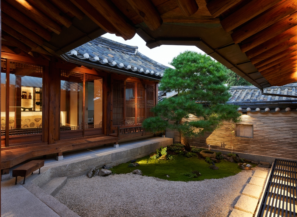 A traditional Korean hanok house in evening with an evergreen tree growing in the centre and big windows showing a few lights inside.