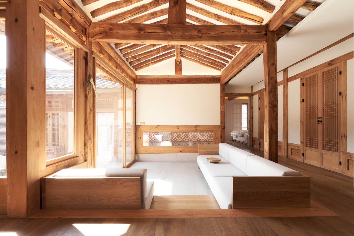 Interior of a living room in a traditional Korean hanok house hotel with exposed beams, white walls and a minmalist white sofa, with floor-to-ceiling windows overlooking a courtyard.