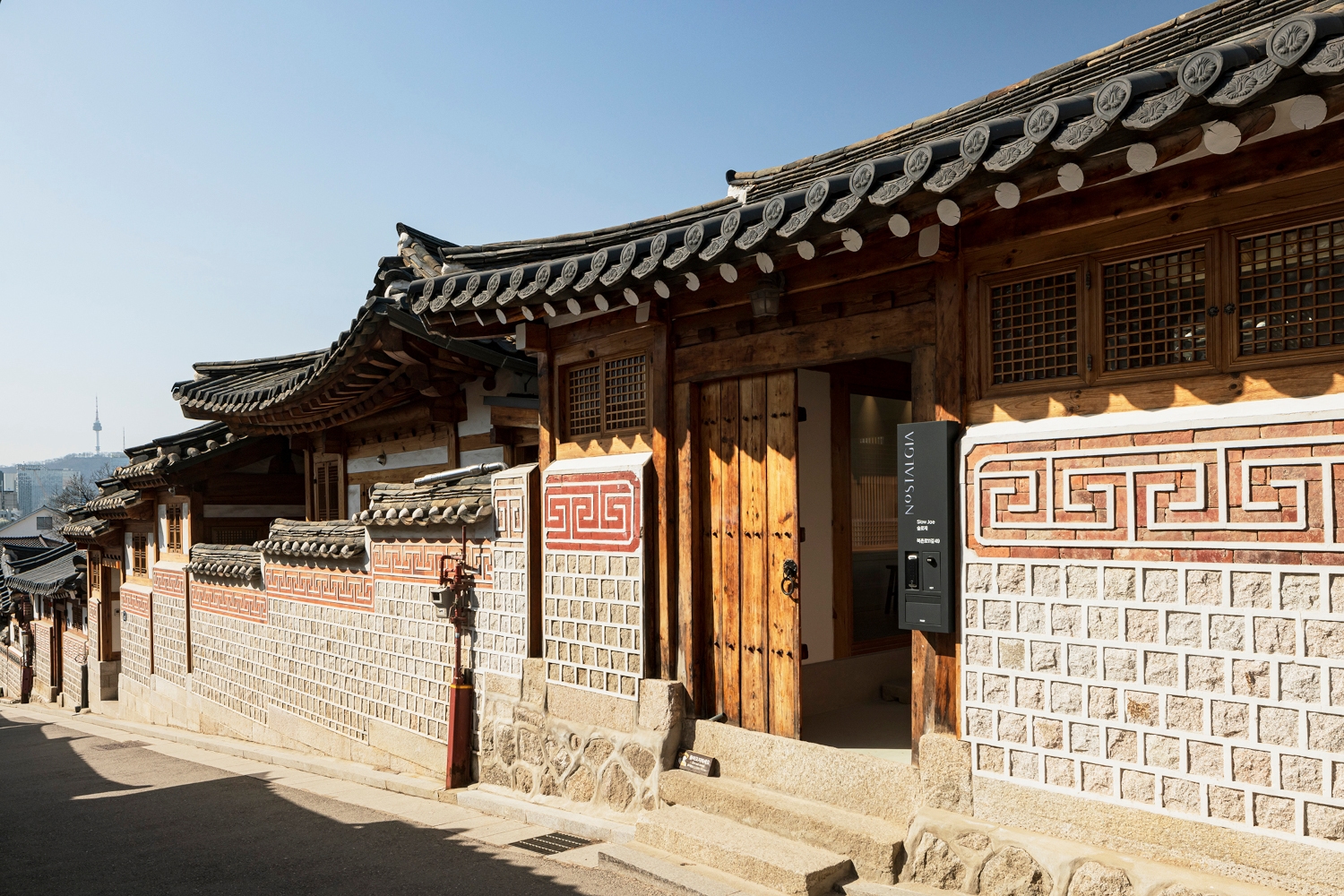 Exterior of the front of a traditional Korean hanok house in an alleyway, with a wooden front door, grey and red tiles, and a traditional tiled roof.