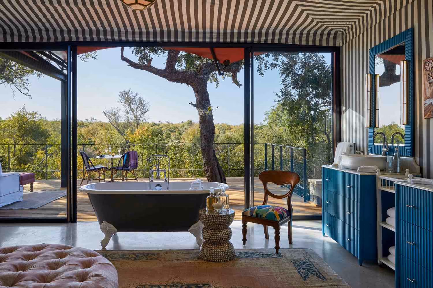A claw-foot bathtub stands in front of a floor to ceiling glass window, leading to a wooden deck and views of the bush. There's a blue fluted vanity and sink unit to the right, and a striped canvas roof above