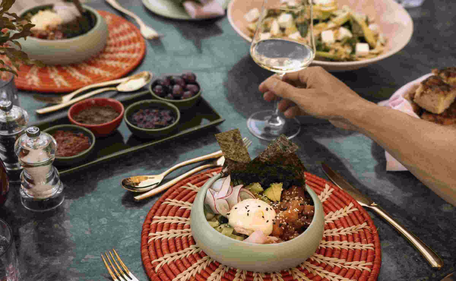 An overhead shot of a marble table laid with bowls of food and condiments, with an arm and hand holding a glass of white wine.