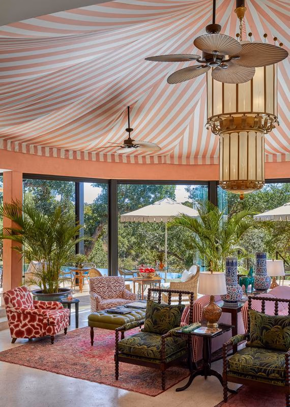 A ceiling fan and glass chandelier hang from a pink and white striped canvas ceiling, above a lounge area with potted palms, patterned armchairs and rugs, with views to the trees outside through tall windows.