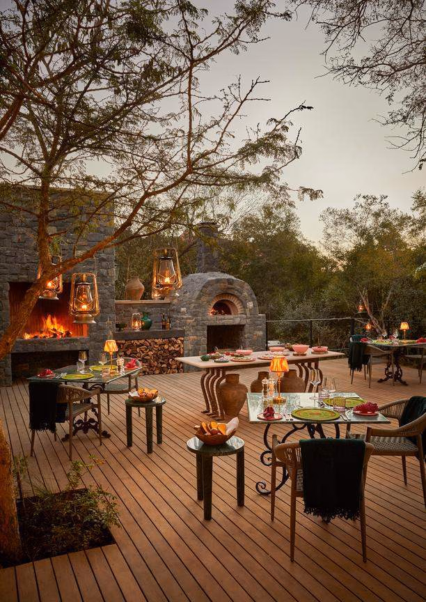 An outdoor area with wooden deck, sevenral tables and chairs and a bigger wooden table laid with food. The light is dusky and there's a tree with lanterns, a fire set in a stone wall and a pizza oven built from the same stone.