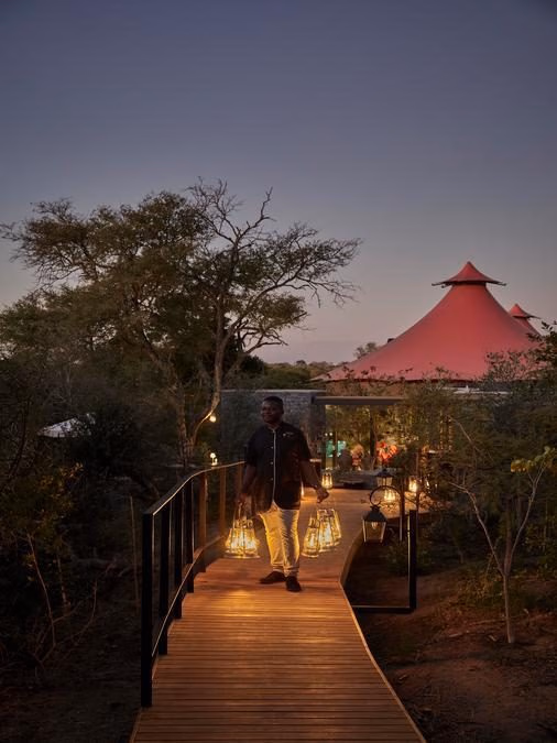 A nighttime scene with a curved wooden walkway leading to red-topped safari tents, with trees at either side. A man in pale trousers and a dark jacket holds several lanterns in each hand.