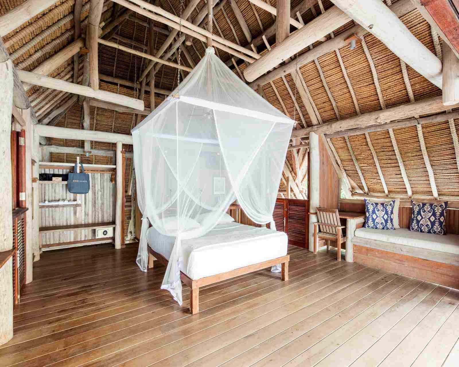 A large, wooden, four-poster bed made up with white linens and draped with sheer white gauze, in a room with wooden floors, thatched walls and roof, and built-in wooden benches