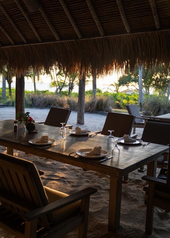 A long table is set for dinner on the beach under a thatched roof, with several chairs and views of the sand, trees and ocean