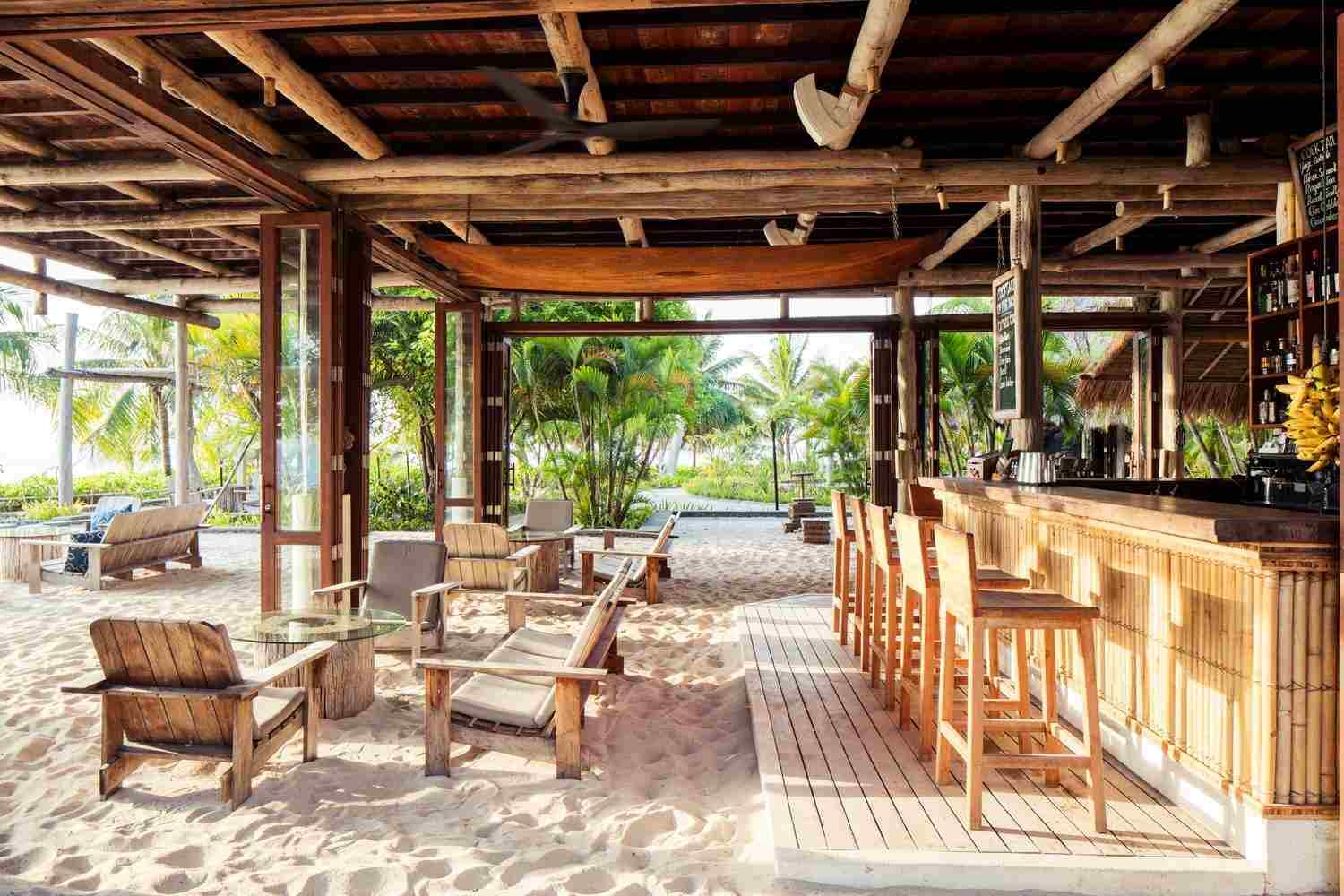 A side view of a beach bar, with wooden chairs and benches in the pale sand, a long, bamboo bar with stools and a wooden roof above, with views of palm trees all around.