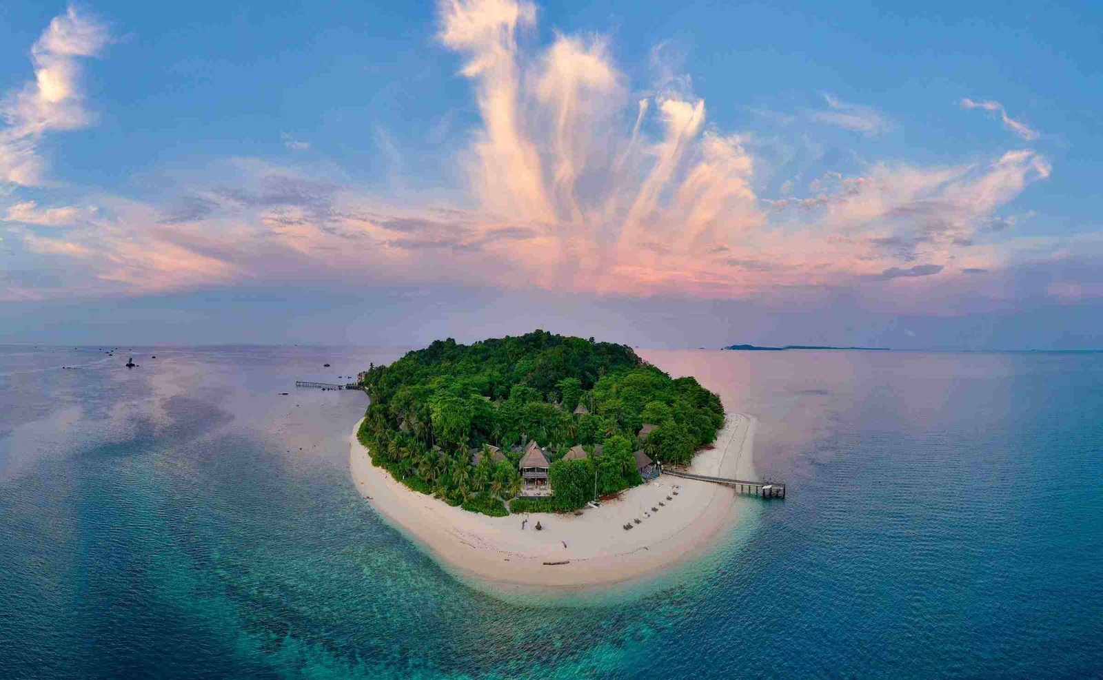 An aerial view of a small island surrounded by the ocean, with pale sandy beaches and a densely forested interior, and a jetty visible to the right. You can see the roofs of villas through the trees.