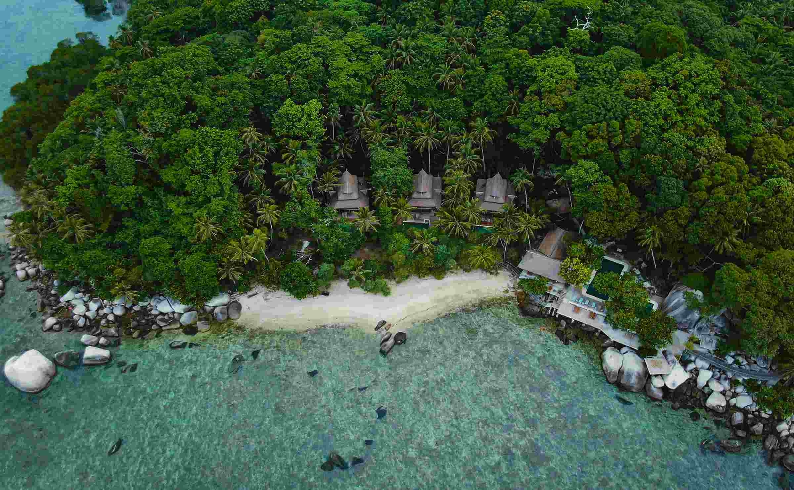 An overhead view of a white-sand beach with dense rainforest and the rooftops of villas visible behind.