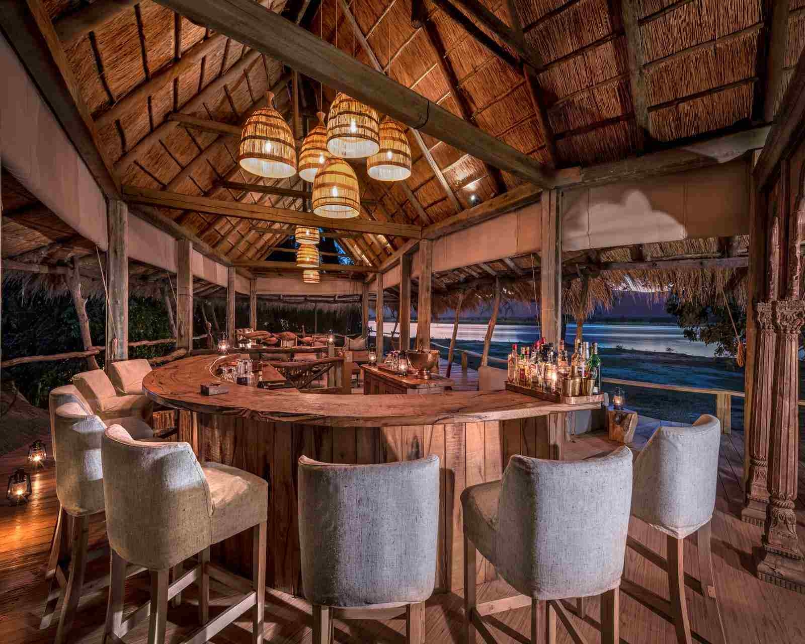 High backed bar stools are set in a semi-circle around a wooden bar, under a thatched palapa roof with nighttime views of the river and mountains behind. Several bottles of spirits can be seen to the side.