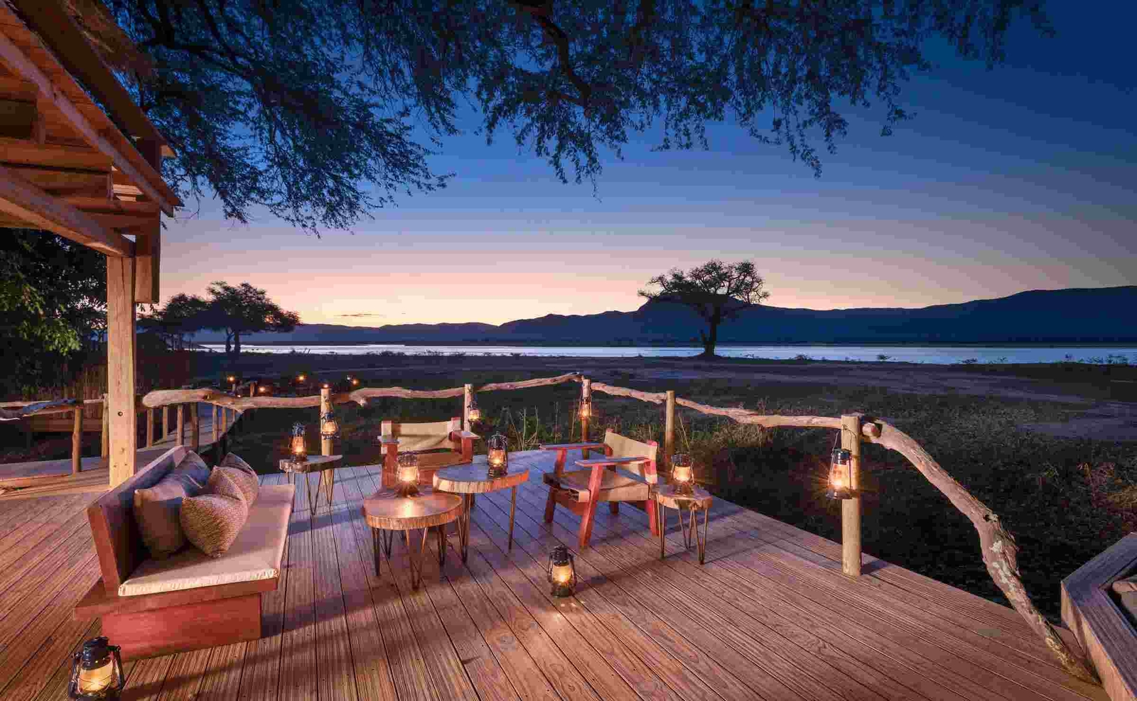 Dusk turning to night skies against a river and mountain backdrop, with a wooden deck set with a sofa, small tables and chairs in the foreground.