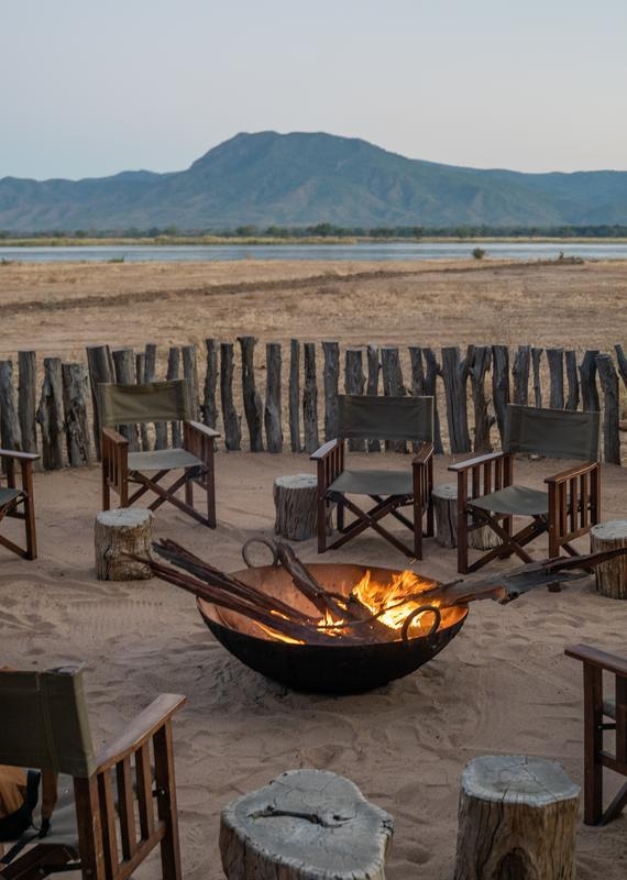 A fire in a bowl firepit is in the centre of a circle of wood and canvas chairs, surrounded by sticks forming another circle, with Rift Valley mountain and Zambezi River views behind