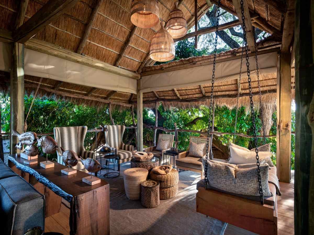 Wooden shelves and armchairs of various shapes of sizes are set around rattan stools under a thatched palapa roof, with views of greenery all around.
