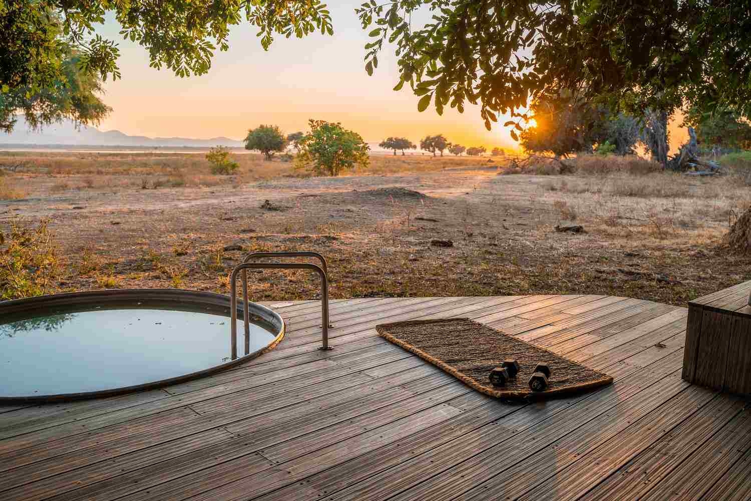 Awooden deck has a rattan mat with two dumbbell weights, with a round plunge pool to the side, accessed by metal steps. There are sunset views of the escarpment and mountains beyond.