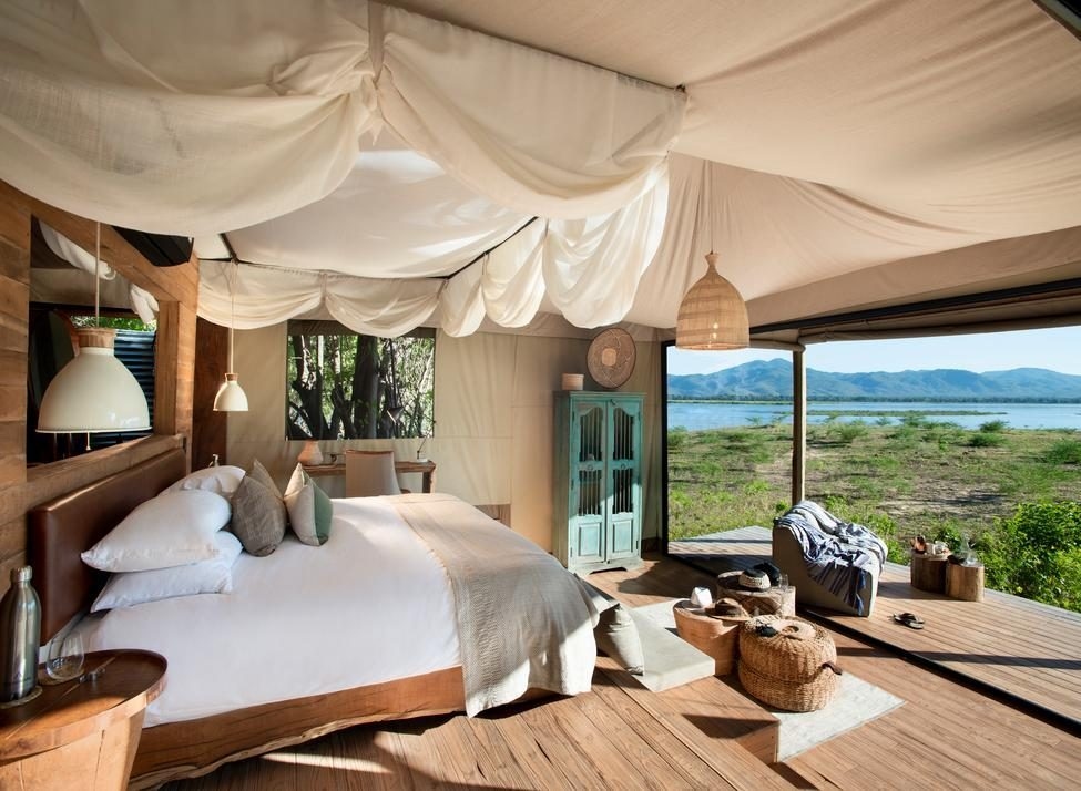 A bed with wooden frame and white linens under canvas on wooden floorboards, looking out to grassland, a river and mountains in the background. The room is furnished with wooden furniture, luxury textiles and rattan seats.