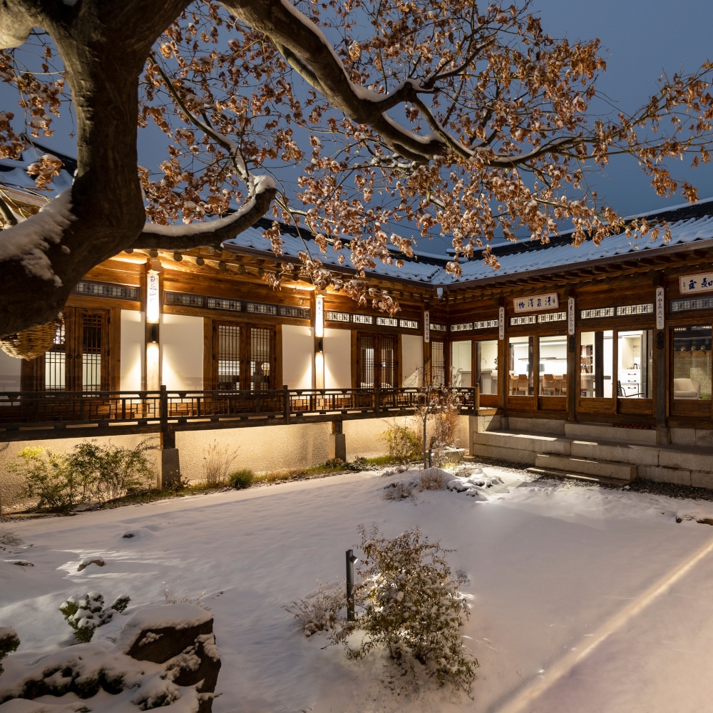 A traditional Korean hanok house on a snowy evening with a tree in the centre and big windows showing a few lights inside.