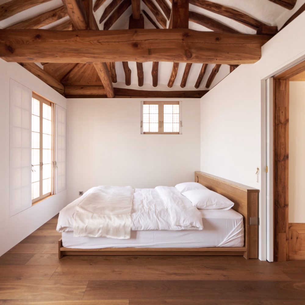Interior of a bedroom in a traditional Korean hanok house hotel with exposed beams, white walls and a minimalist white bed, with lattice windows allowing in bright light.