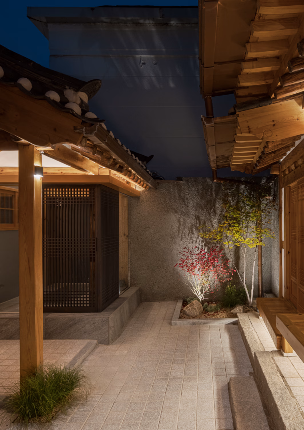 A traditional Korean hanok house in evening with a small tree growing in the corner and big windows showing a few lights inside.