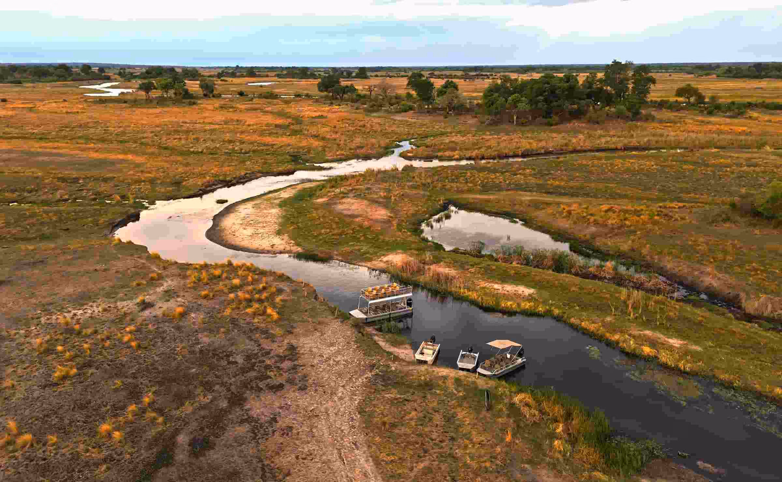 An overhead view of the wetlands, with one large and three smaller boats seen on the edge of a waterway, and patches of forest in the background.