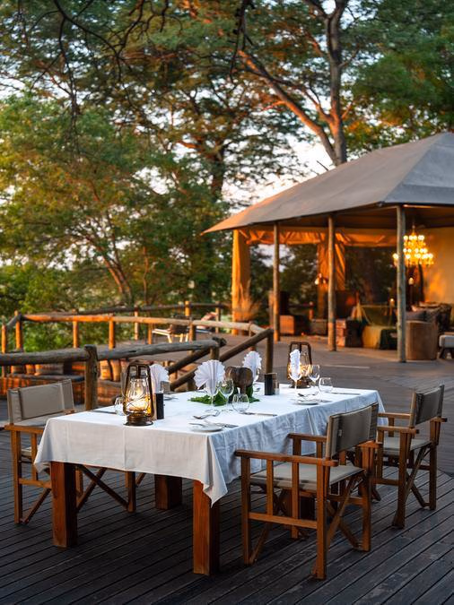 A table set for four people is in the foreground on a wooden deck, with a tent and trees in the background.